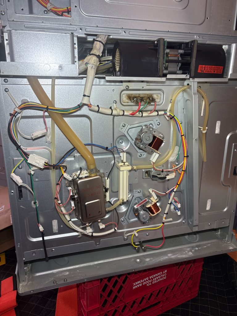 Samsung NQ70M7770DS combo oven pulled from its wall cabinet and set on a protective blanket and milk crate, rear panel off to expose the steam holder assembly and wiring during a South Calgary repair